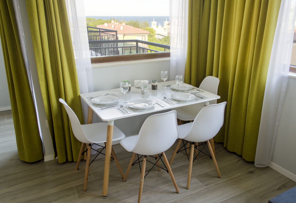 Dining table with modern white chairs and lime green curtains framing sea view